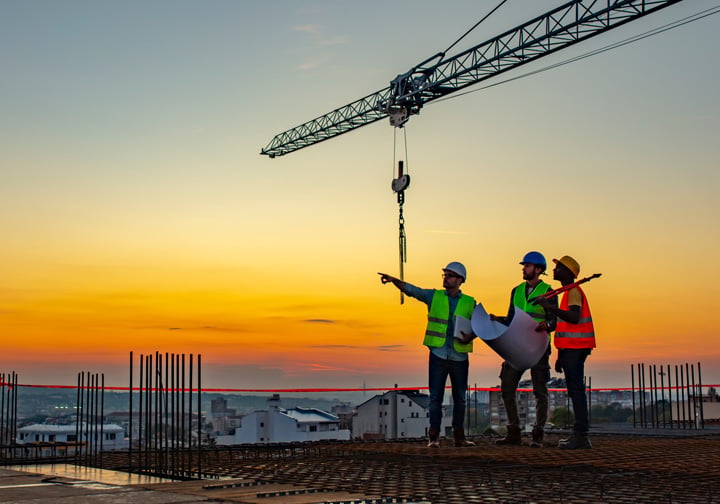 Three people stand on top of a building wearing safety gear, with a crane and an orange sky in the background. 