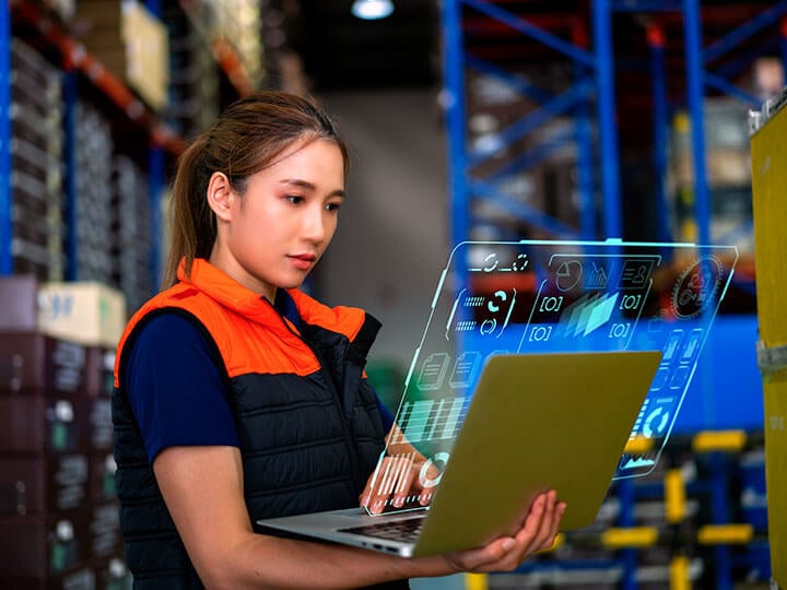 Asian female warehouse worker, in orange and black vest, looking on a laptop with a cybersecurity concept displayed 