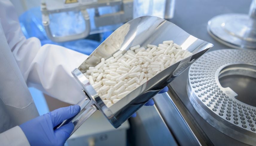 Gloved technician holding a scoop of white capsules in a pharmaceutical manufacturing facility.